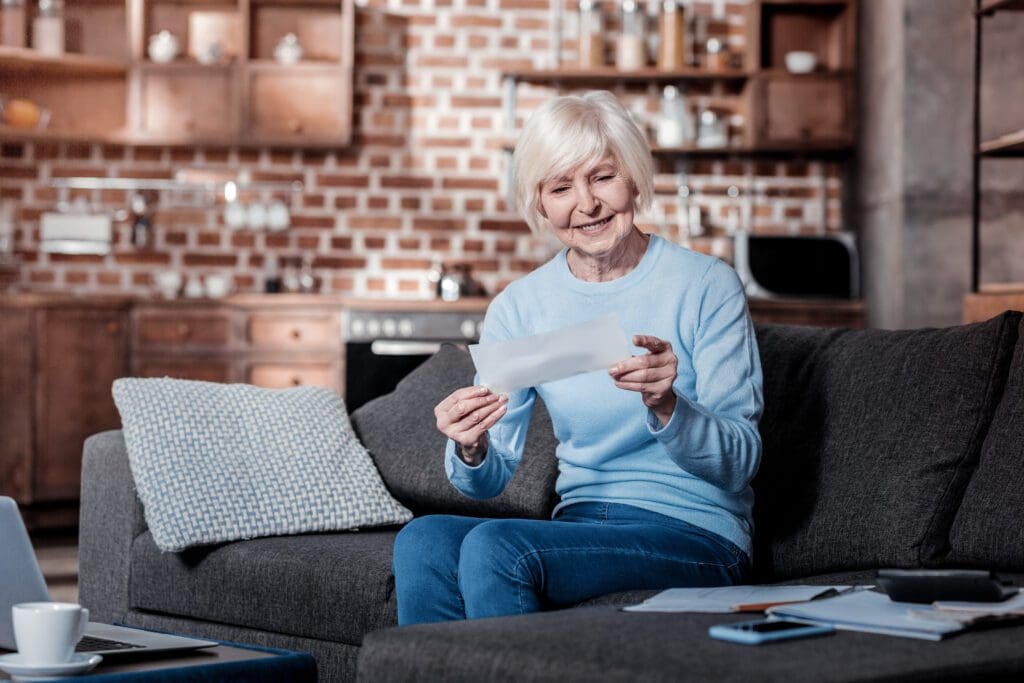 Smiling elderly woman sitting on a sofa in a cozy living room, holding and looking at a piece of paper—possibly related to Social Security—with documents and a mug on the table nearby.