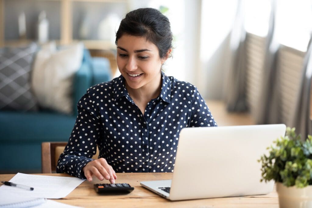 A woman in a navy polka-dot shirt smiles while using a calculator at a desk with a laptop, papers, and a plant, focused on savings and investing as she sits in a brightly lit living room.
