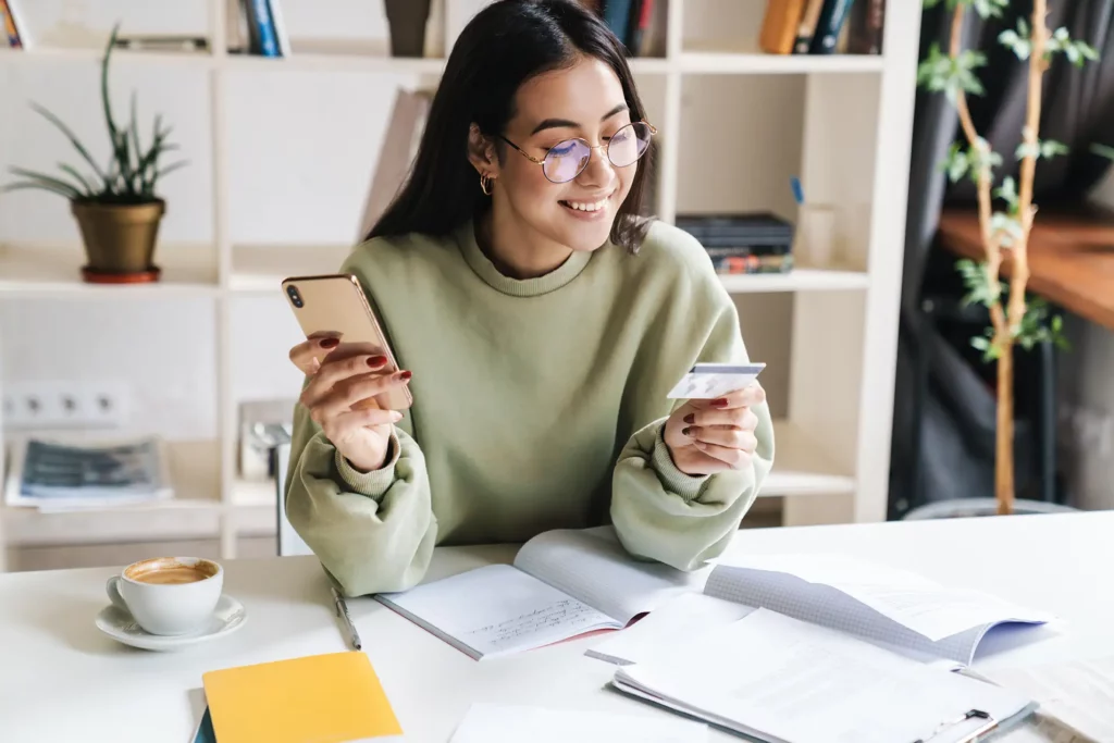 A woman with glasses sits at a desk with books and papers, holding a smartphone in one hand and a credit card in the other, smiling. A cup of coffee and a shelf with plants are in the background.