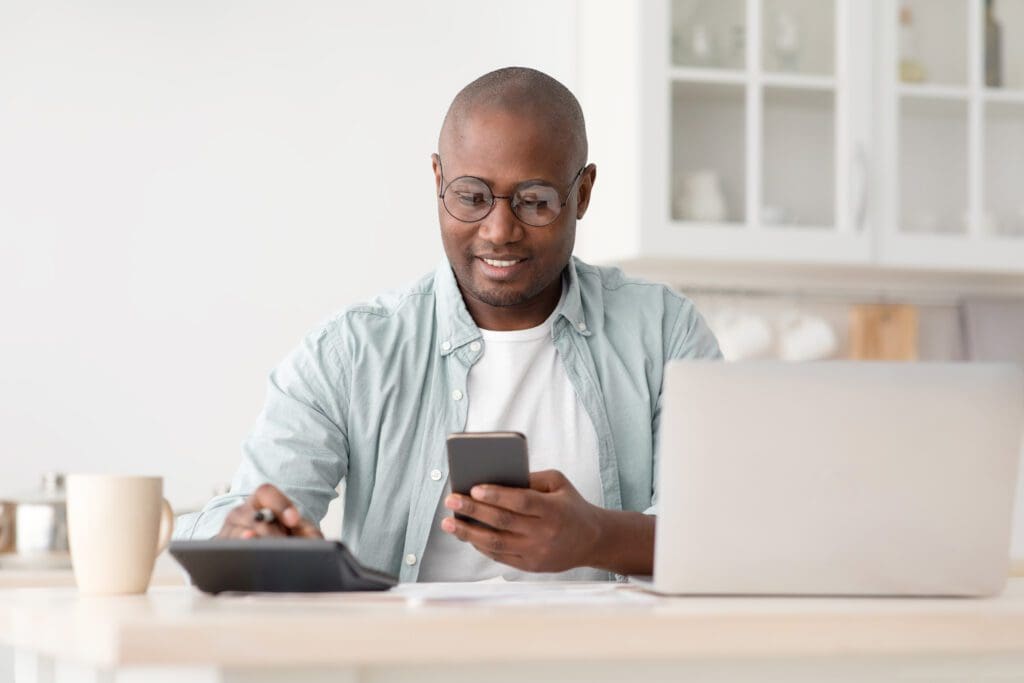 A man sits in front of a computer, holding a phone and a calculator as he calculates his investments.