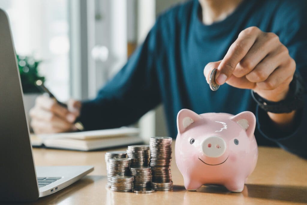 A person makes smart money moves by dropping a coin into a pink piggy bank beside stacks of coins, with a laptop and notebook on the desk in a bright room.