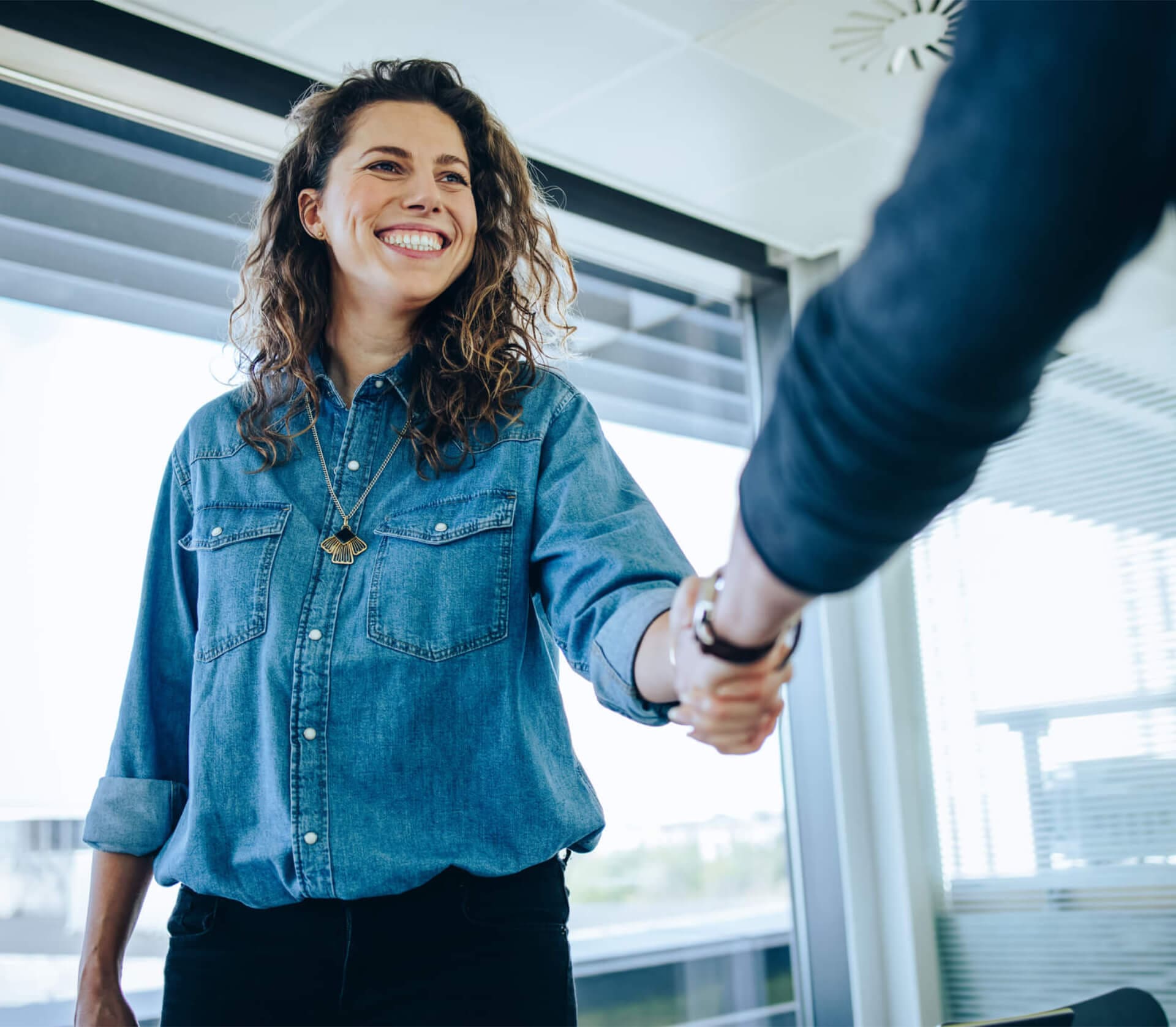 A woman in a denim shirt smiles while shaking hands with another person in an office setting.