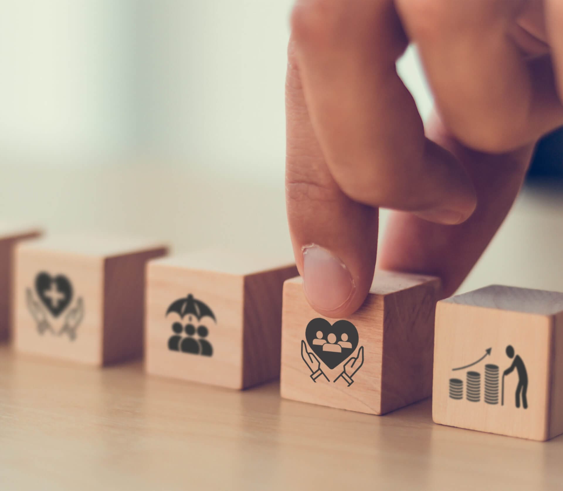 A hand selects a wooden block with an icon of a heart and people inside, symbolizing care or wellbeing. Other blocks show icons for health, insurance, and growth.
