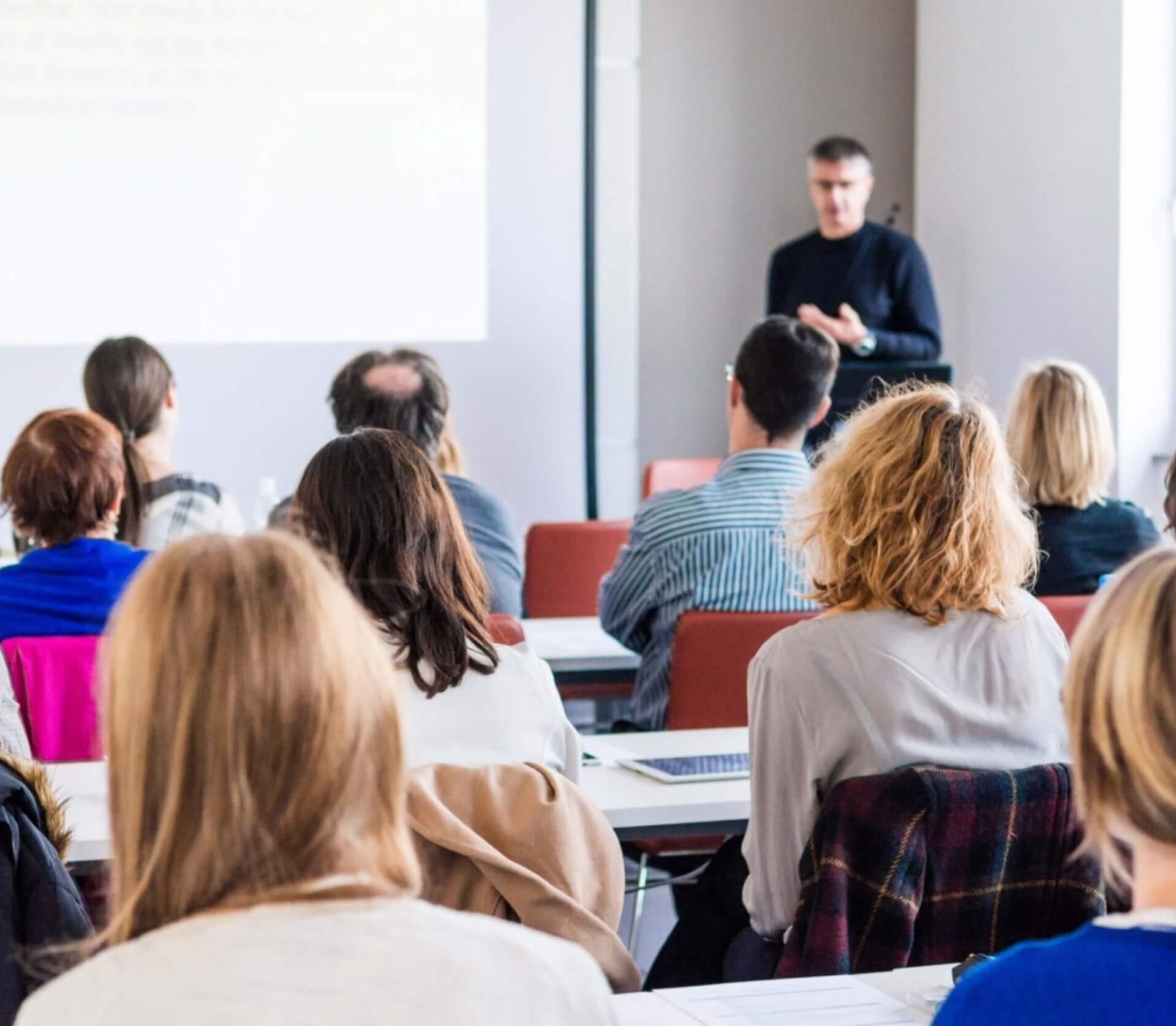 A group of people sit at desks facing a speaker who is presenting in front of a screen in a bright classroom or seminar room.
