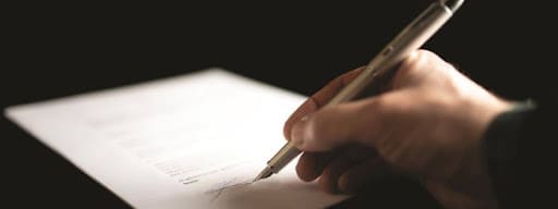 A close-up of a hand holding a pen and signing a document on a white sheet of paper, with a dark background.